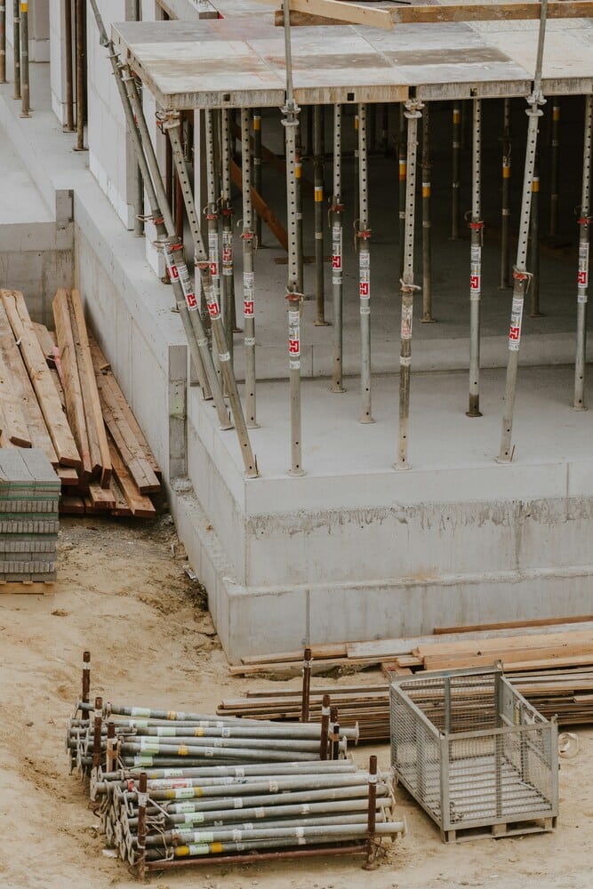 An elevated view of a construction site showing adjustable steel shoring props supporting a multi-level concrete foundation structure next to building materials.