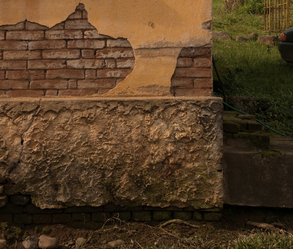 Severe damage to an exterior brick foundation showing cracked plaster, crumbling concrete, and exposed masonry.