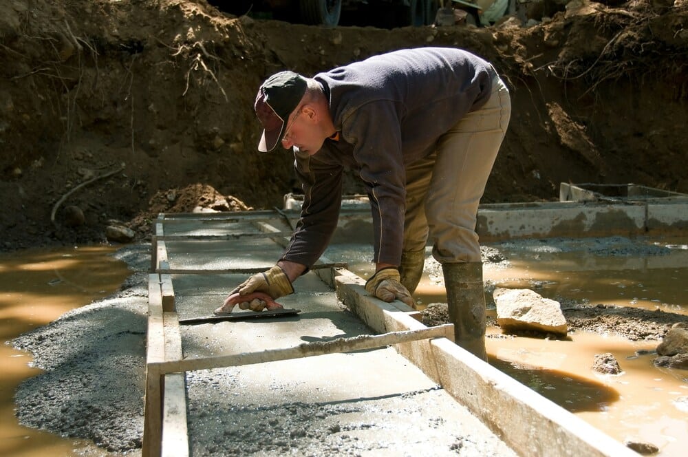 A construction worker wearing a cap, gloves, and tall rubber boots stands in a muddy trench alongside a house, carefully leveling out a freshly poured concrete footing strip with a hand trowel tool.