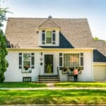 Front view of a white, well-maintained two-story suburban house with dark window shutters on a sunny day. A homeowner stands by the front window boxes adjusting flower pots, demonstrating the care taken to protect landscaping before a repair crew arrives.