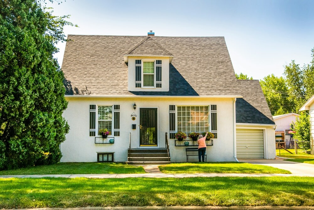 Front view of a white, well-maintained two-story suburban house with dark window shutters on a sunny day. A homeowner stands by the front window boxes adjusting flower pots, demonstrating the care taken to protect landscaping before a repair crew arrives.