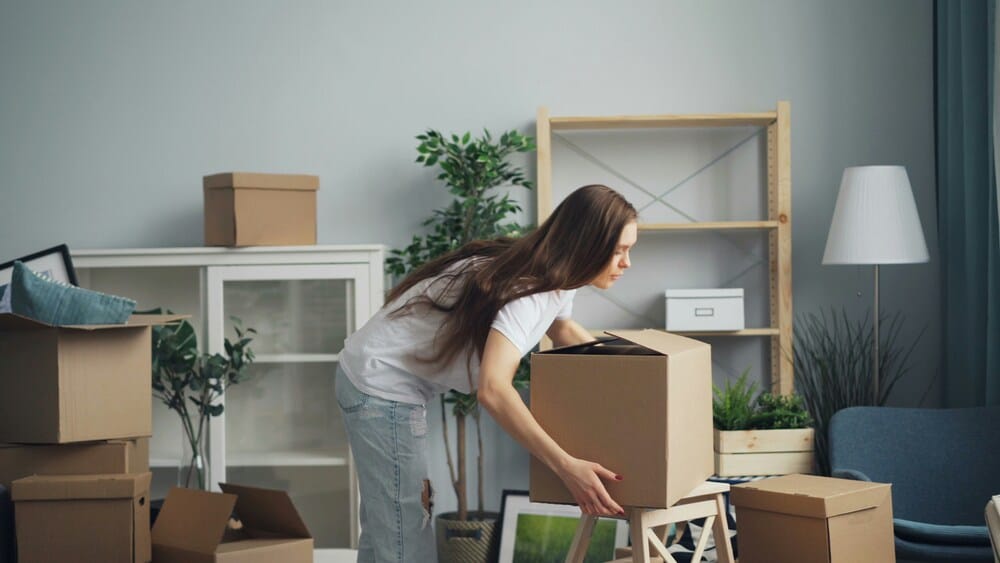 A woman inside her living room carefully packing books and breakables into a large cardboard box, preparing her home's interior and clearing items from shelves to protect them from structural vibrations.