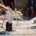 Construction worker hosing water across a rebar-reinforced concrete slab while a crew works, keeping the pour moist as it cures.