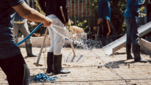 Construction worker hosing water across a rebar-reinforced concrete slab while a crew works, keeping the pour moist as it cures.